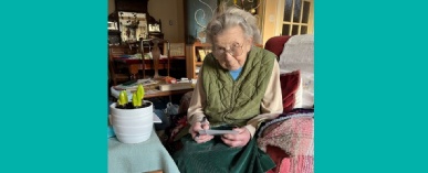 Miss Rosemary Tice seated in an arm chair, writing a greetings card, with the Stephen Perse Cambridge birthday hyacinths on the desk beside her.
